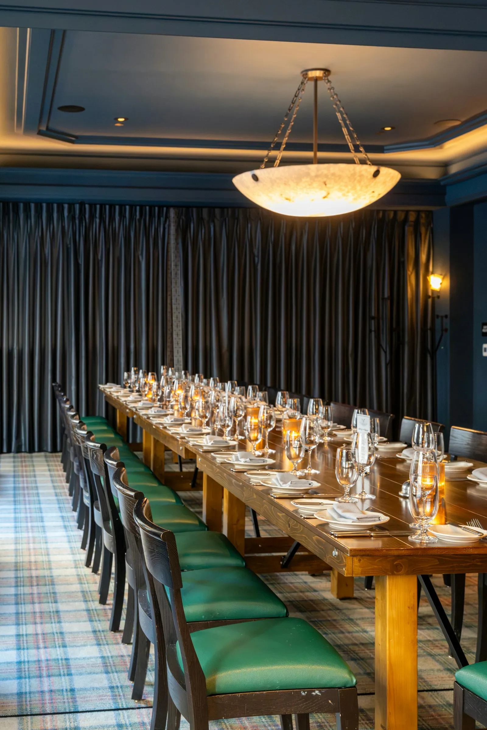 A long wooden table set for dinner in a private room with green chairs and a pendant lamp