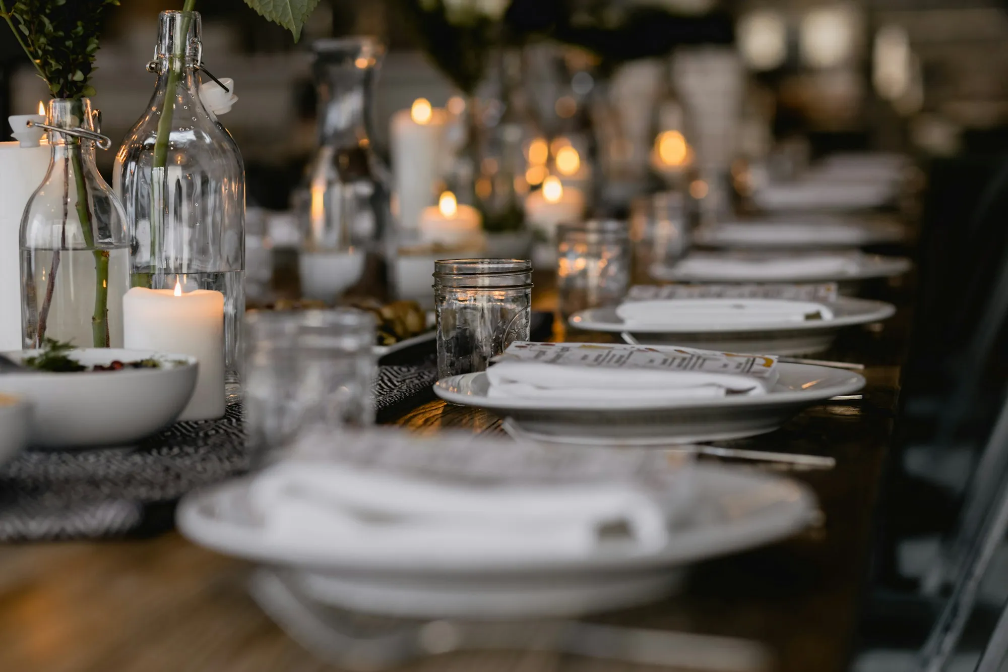 A long table set for a dinner party with candles, glasses and white plates
