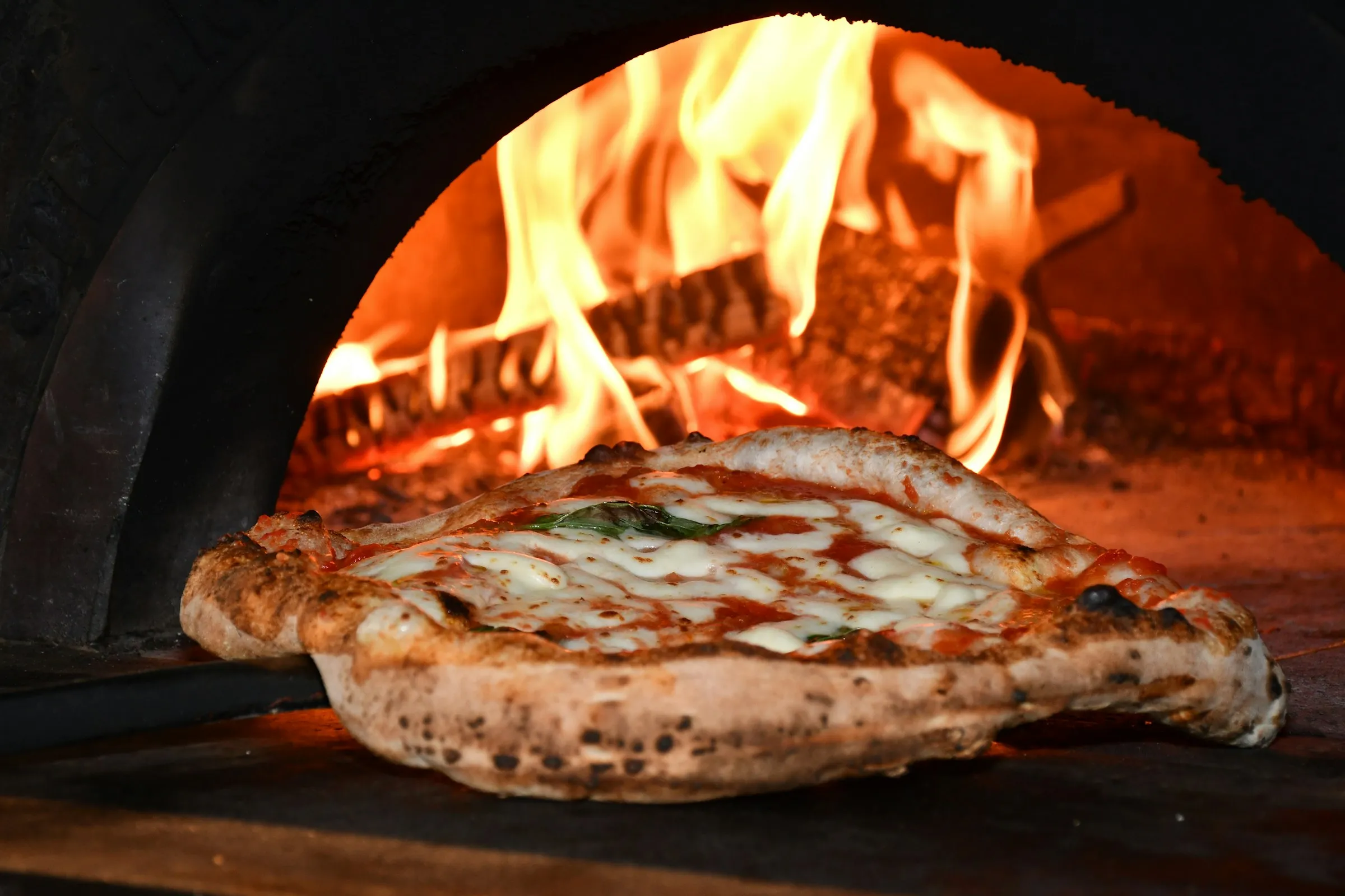 A flatbread baking on the floor of the wood-fired oven, flames curling over the dome
